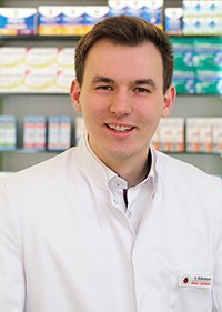 Apotheker Stefan Büllesbach lächelt in die Kamera. Er trägt einen weißen Apothekerkittel und steht vor einem Regal mit verschiedenen Arzneimittel